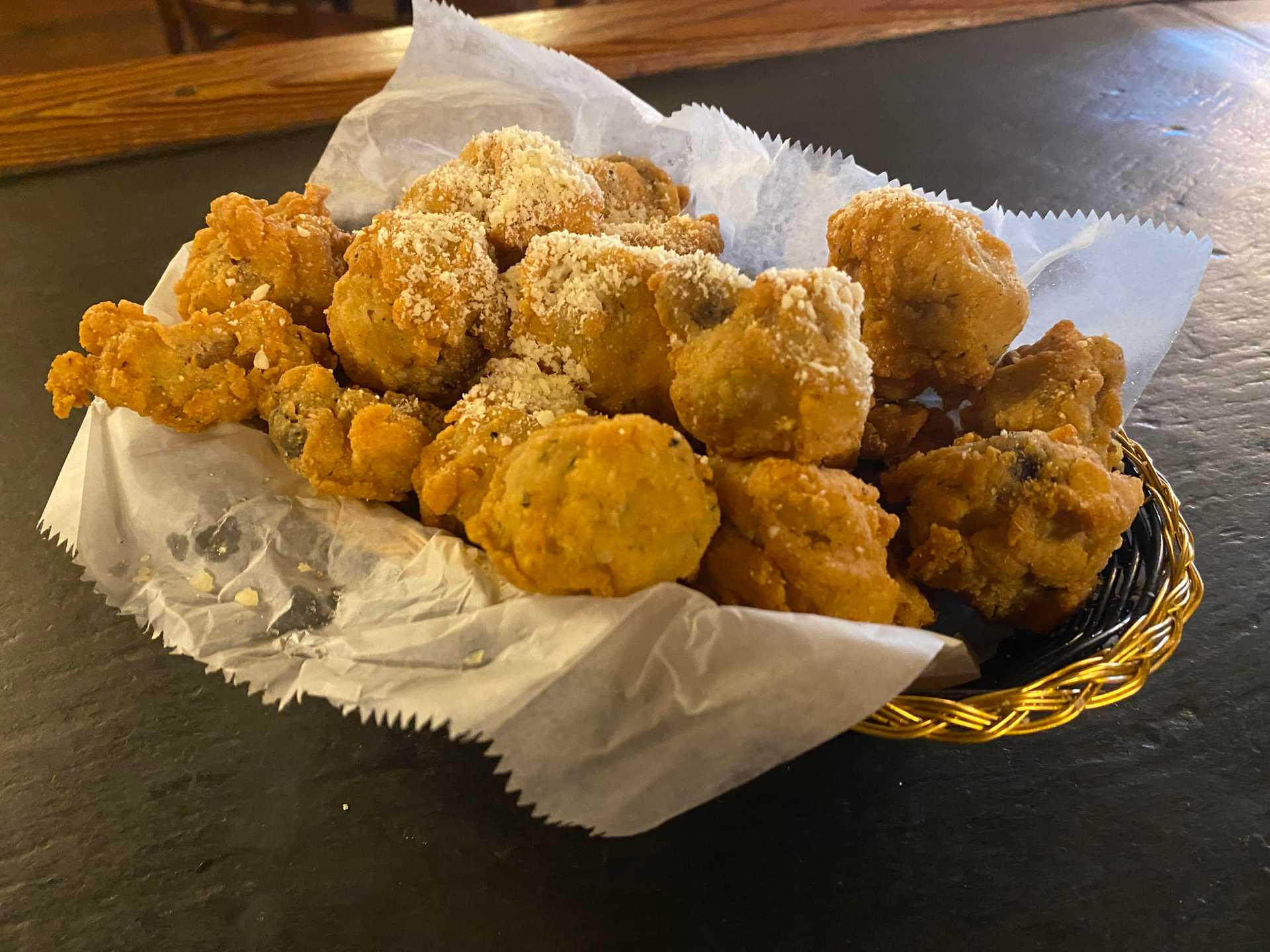 Basket of golden-brown fried mushrooms topped with grated cheese on a wax paper lining.