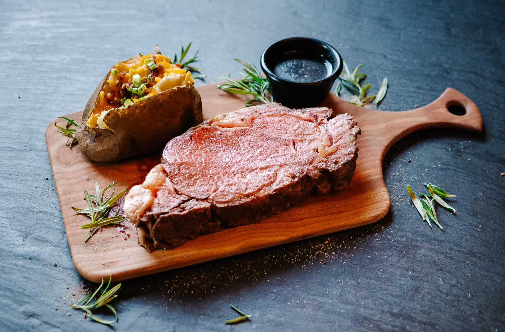 Grilled steak and baked potato with herbs on a wooden cutting board.